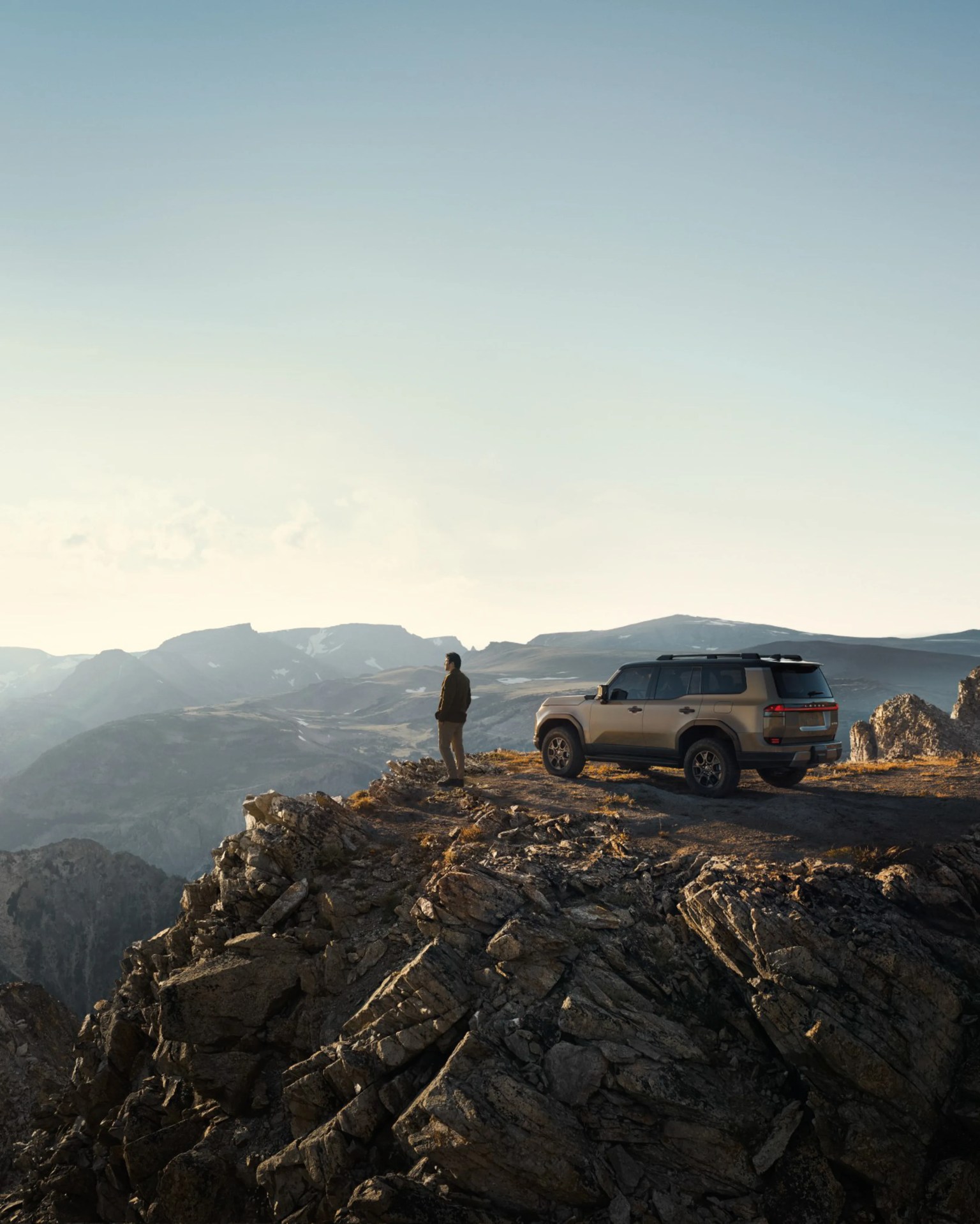 Beige SUV parked on rocky mountain edge with person standing nearby overlooking distant peaks.