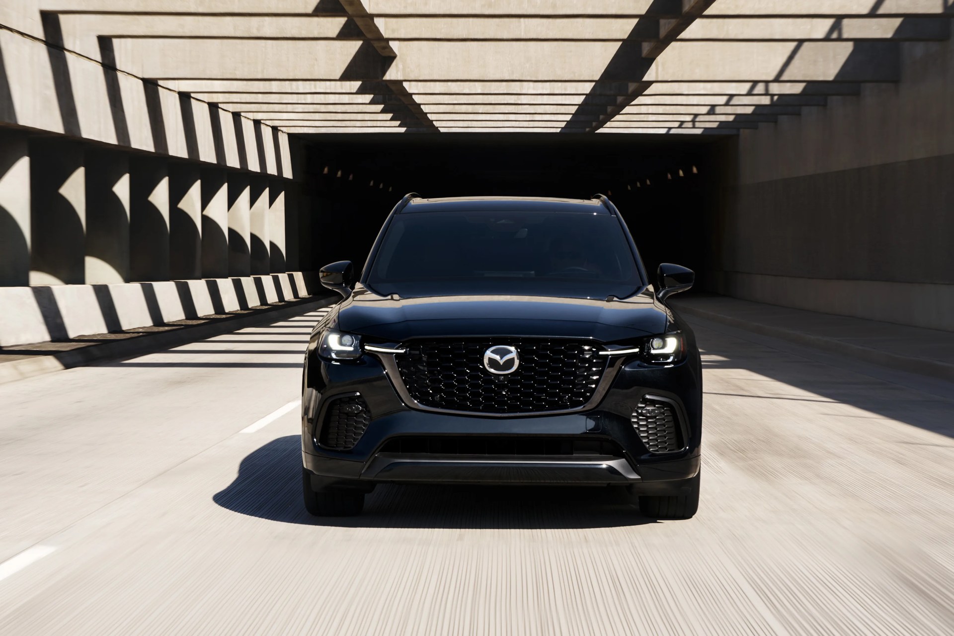 Front view of a black Mazda SUV with headlights on, driving through a tunnel with patterned shadows.