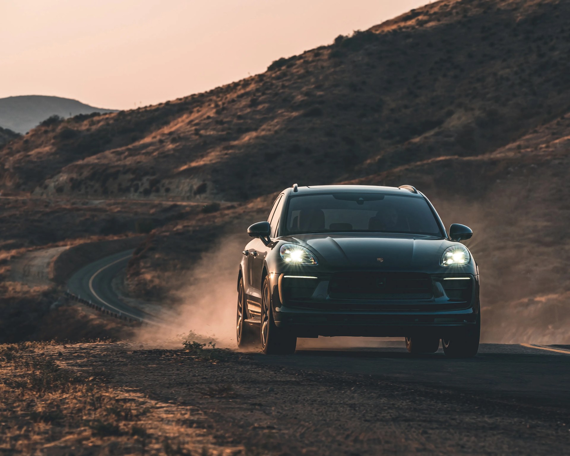 Black Porsche SUV driving on a dusty winding road with hills in the background at sunset.
