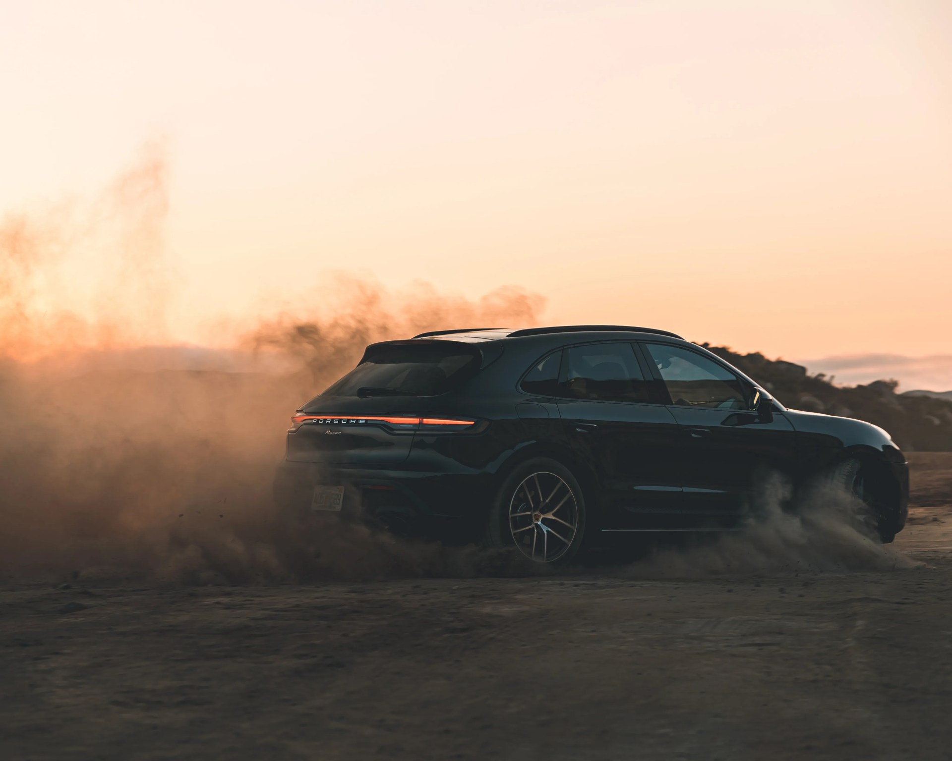 Black Porsche Macan SUV kicking up dust on a dirt road at sunset.