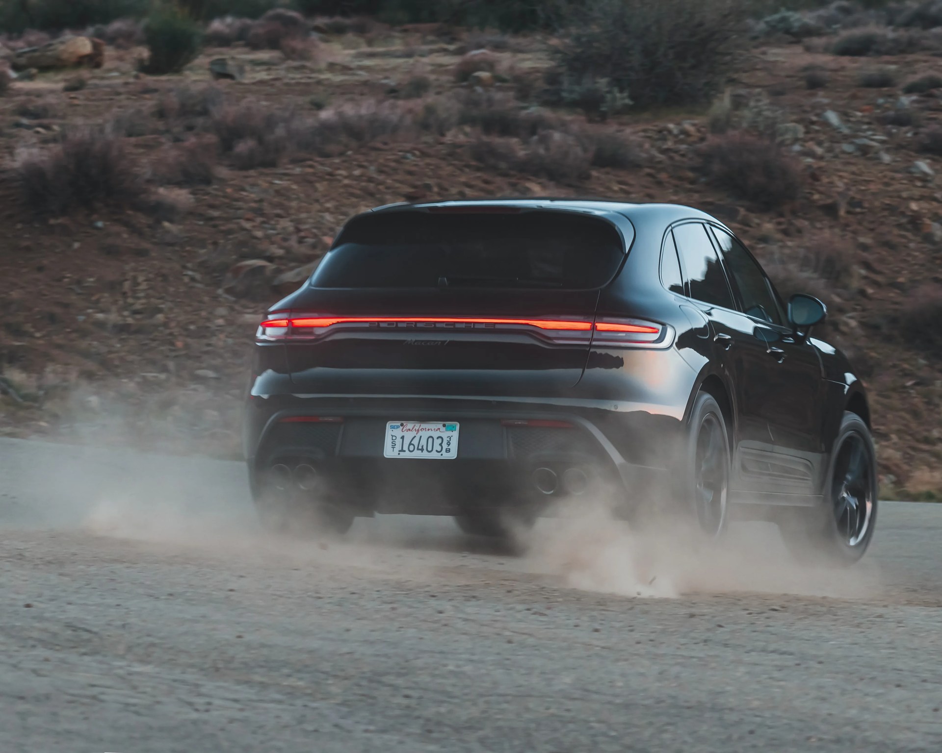 Black Porsche Macan driving on a dusty dirt road with red rear lights illuminated.