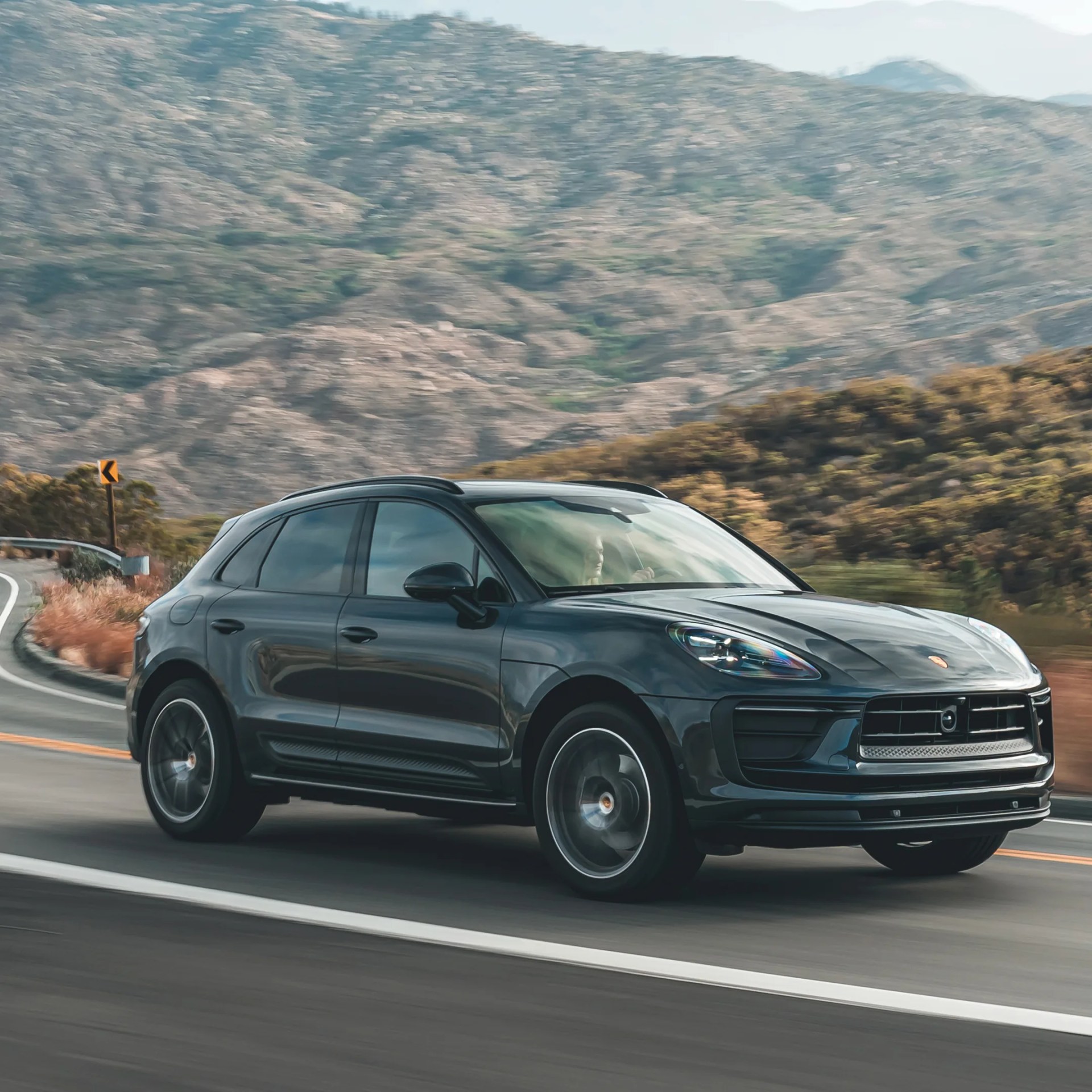 Black Porsche SUV driving on a mountain road with rocky hills in the background.