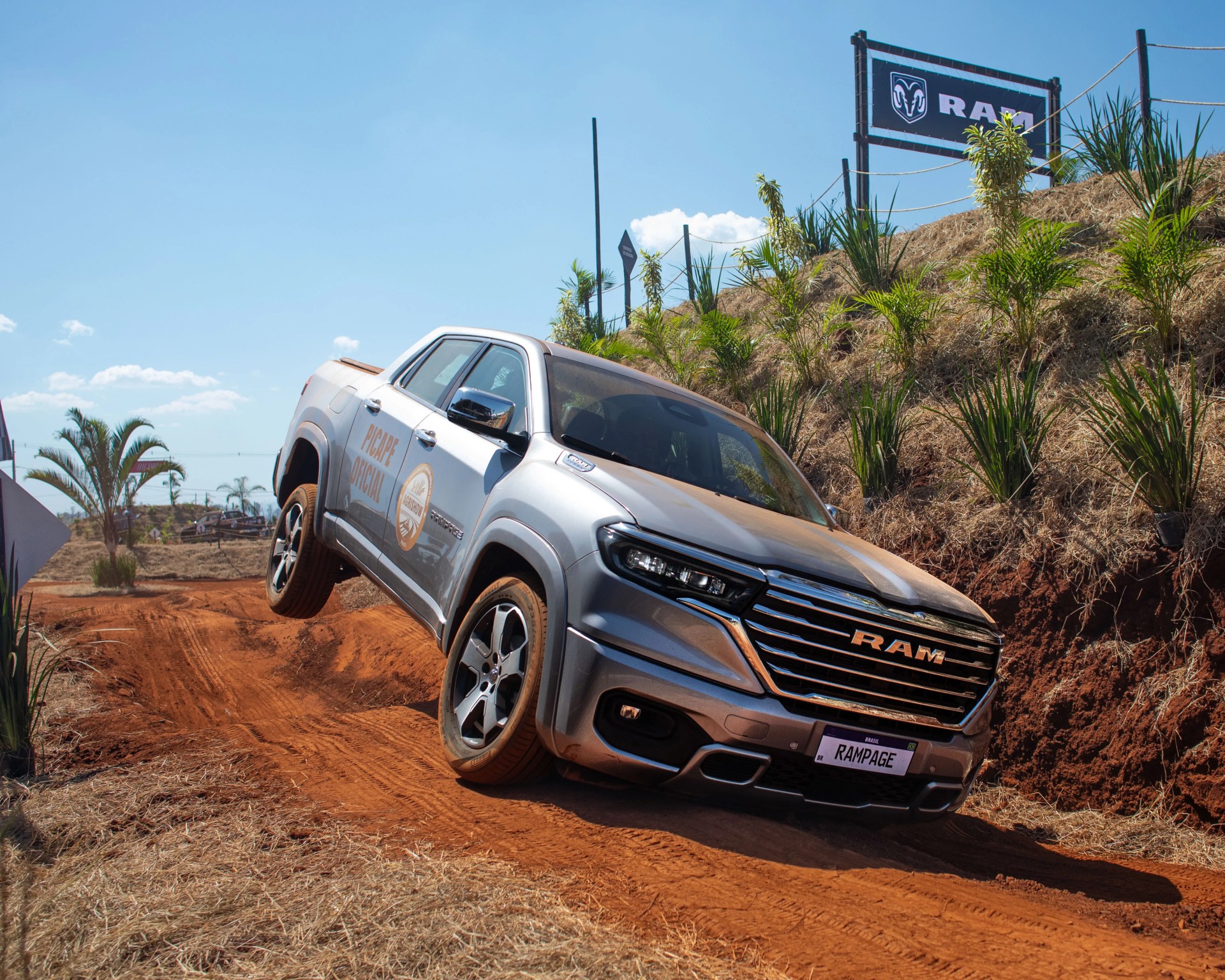 Silver RAM pickup truck driving on a dirt off-road trail with a RAM sign in the background.