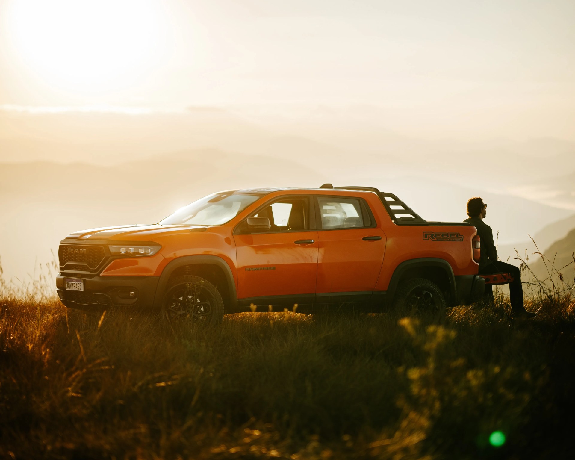 Orange RAM Rebel pickup truck parked in a grassy field at sunset with a person sitting on the tailgate.