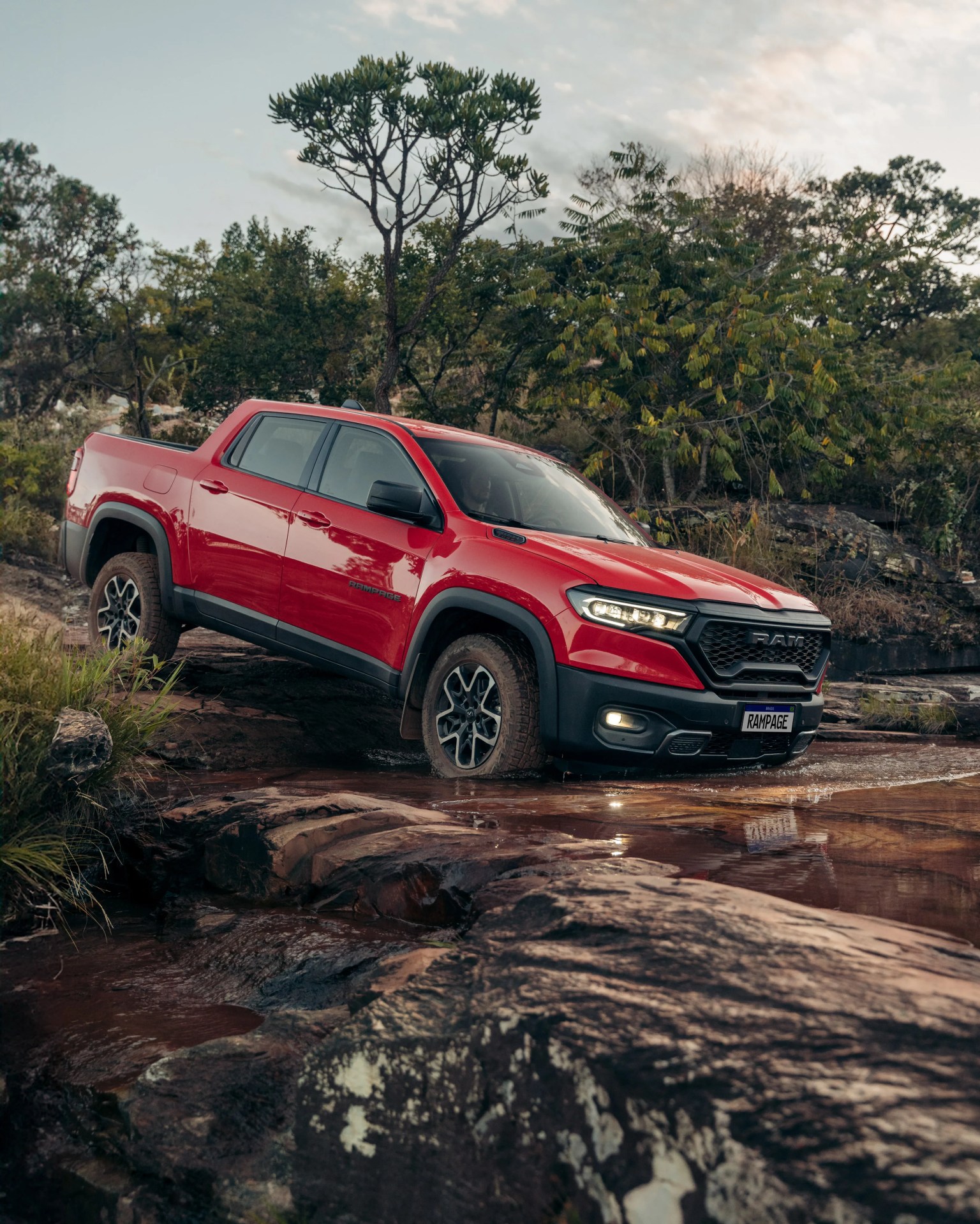 Red RAM Rampage pickup truck driving off-road over rocky terrain with trees in the background.
