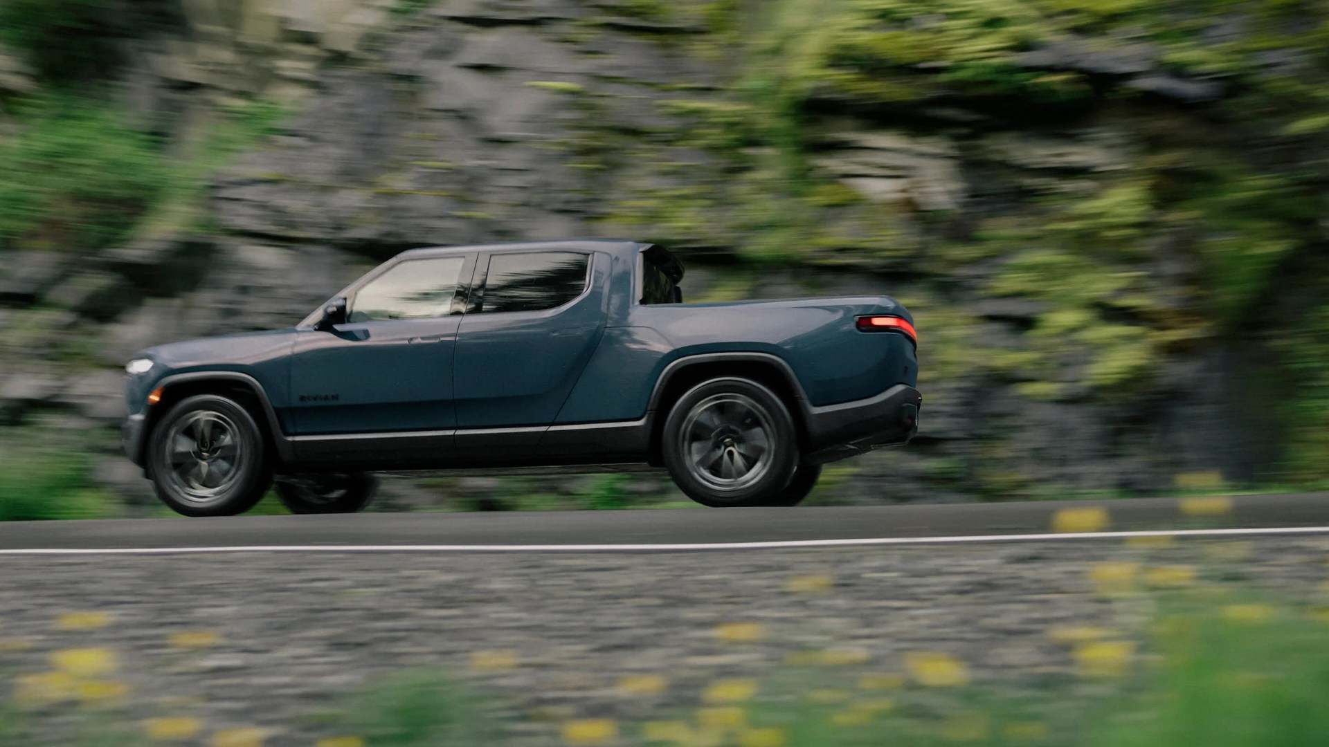 Blue Rivian pickup truck driving on a road with blurred rocky and green background.