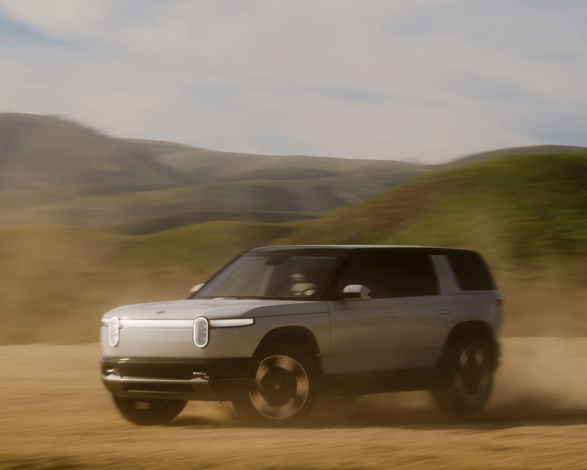 White electric SUV driving on a dusty dirt road with green hills in the background.