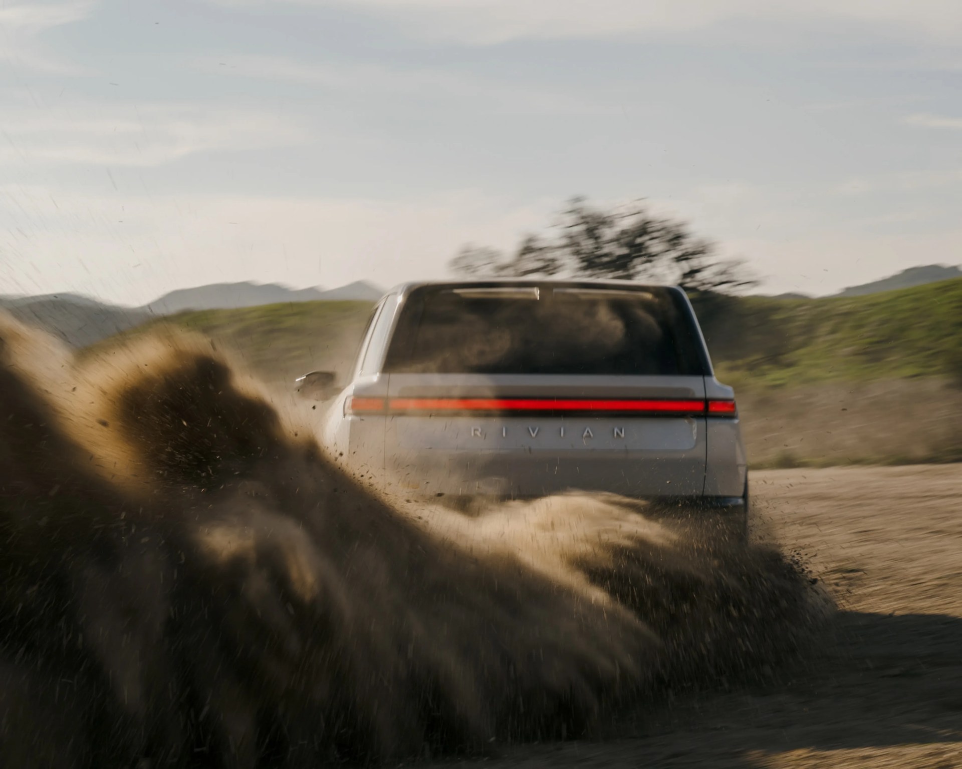 Silver Rivian SUV kicking up dirt on a rural road with hills and trees in the background.