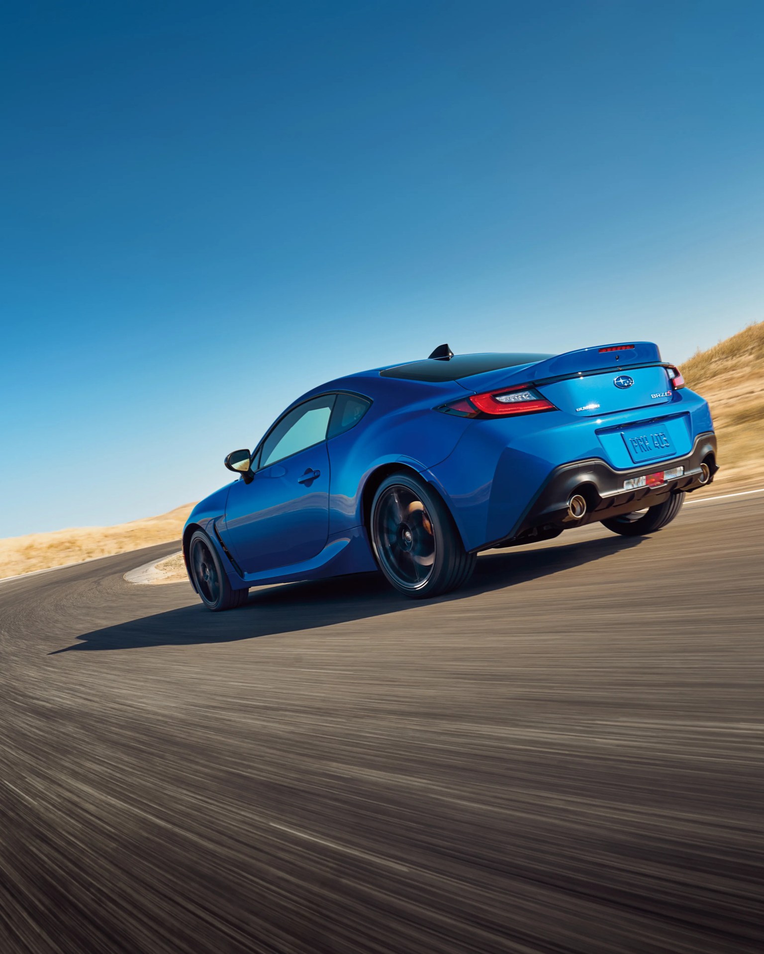 Blue Subaru BRZ sports car cornering on a racetrack under a clear blue sky.
