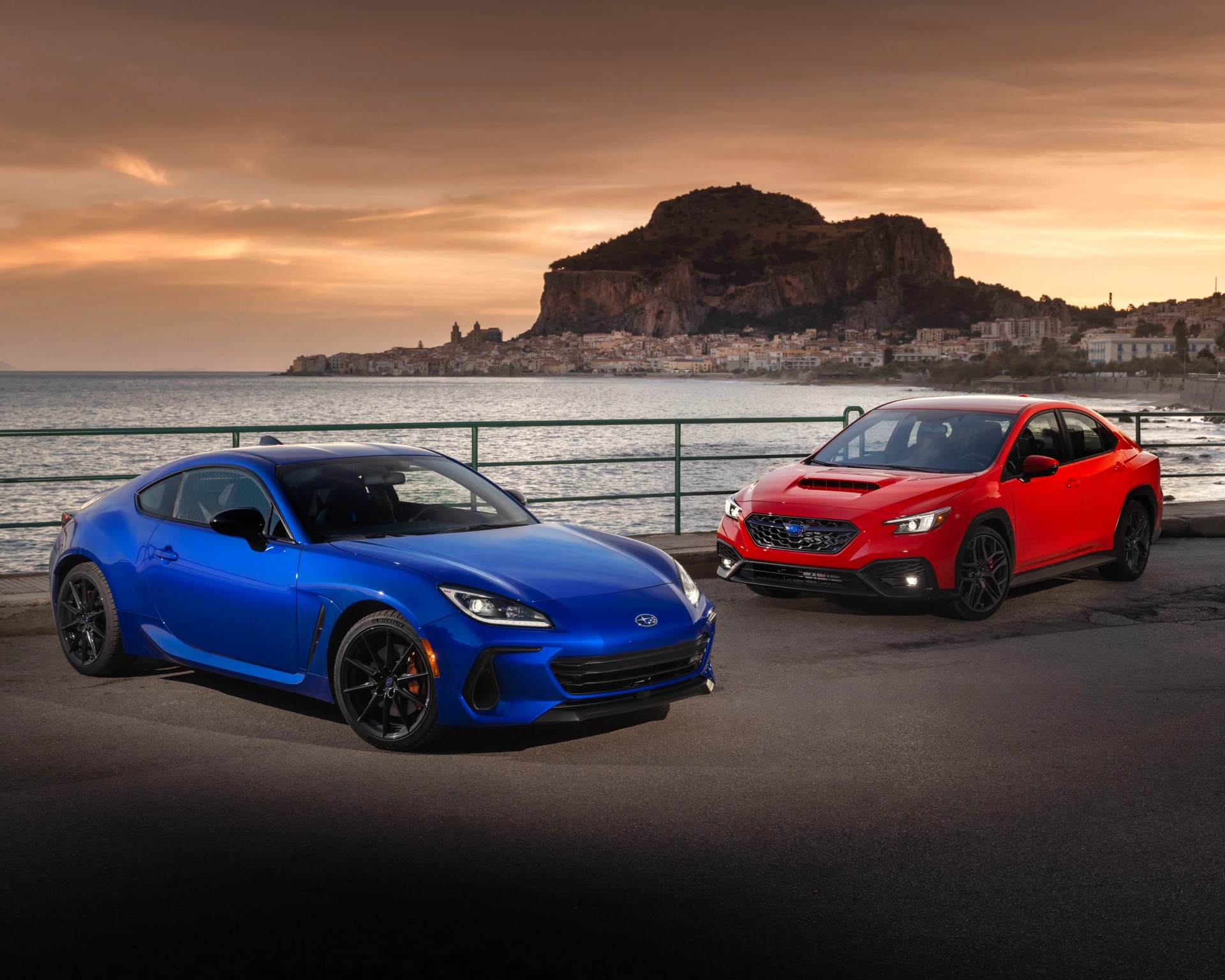 Blue Subaru sports car and red Subaru sedan parked by a waterfront with a mountainous town in the background at sunset.