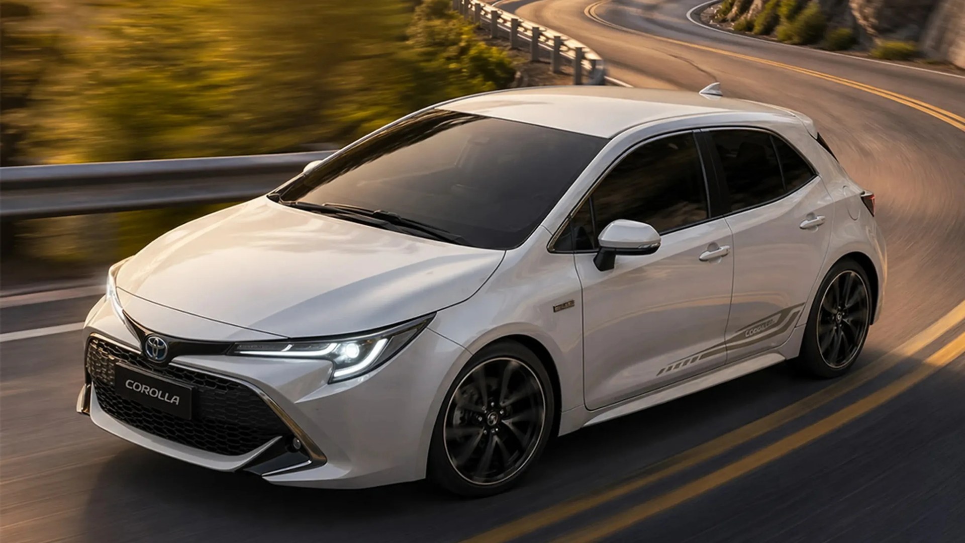 White Toyota Corolla hatchback with black wheels driving on a winding road with guardrails.