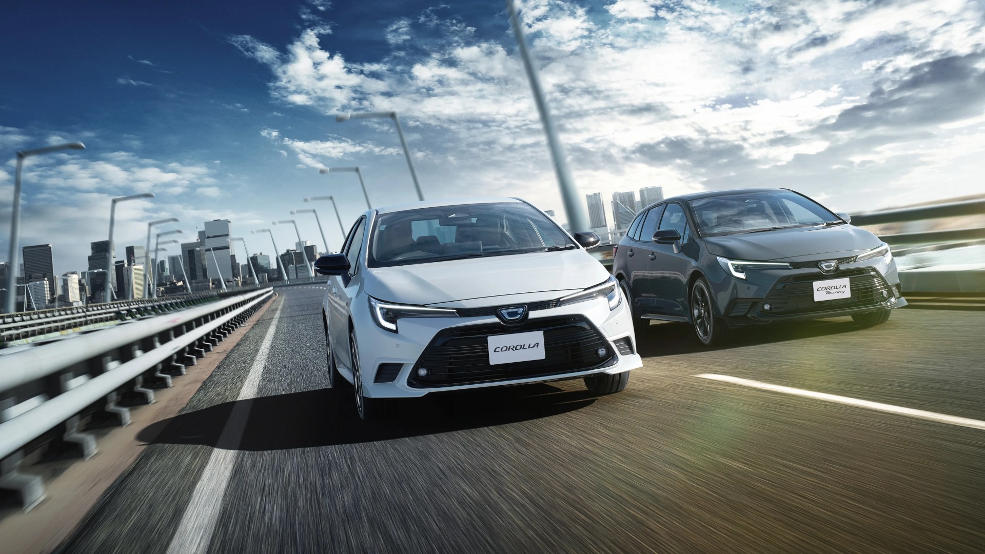 Two Toyota Corolla cars, one white and one dark gray, driving side by side on a highway with a city skyline in the background.