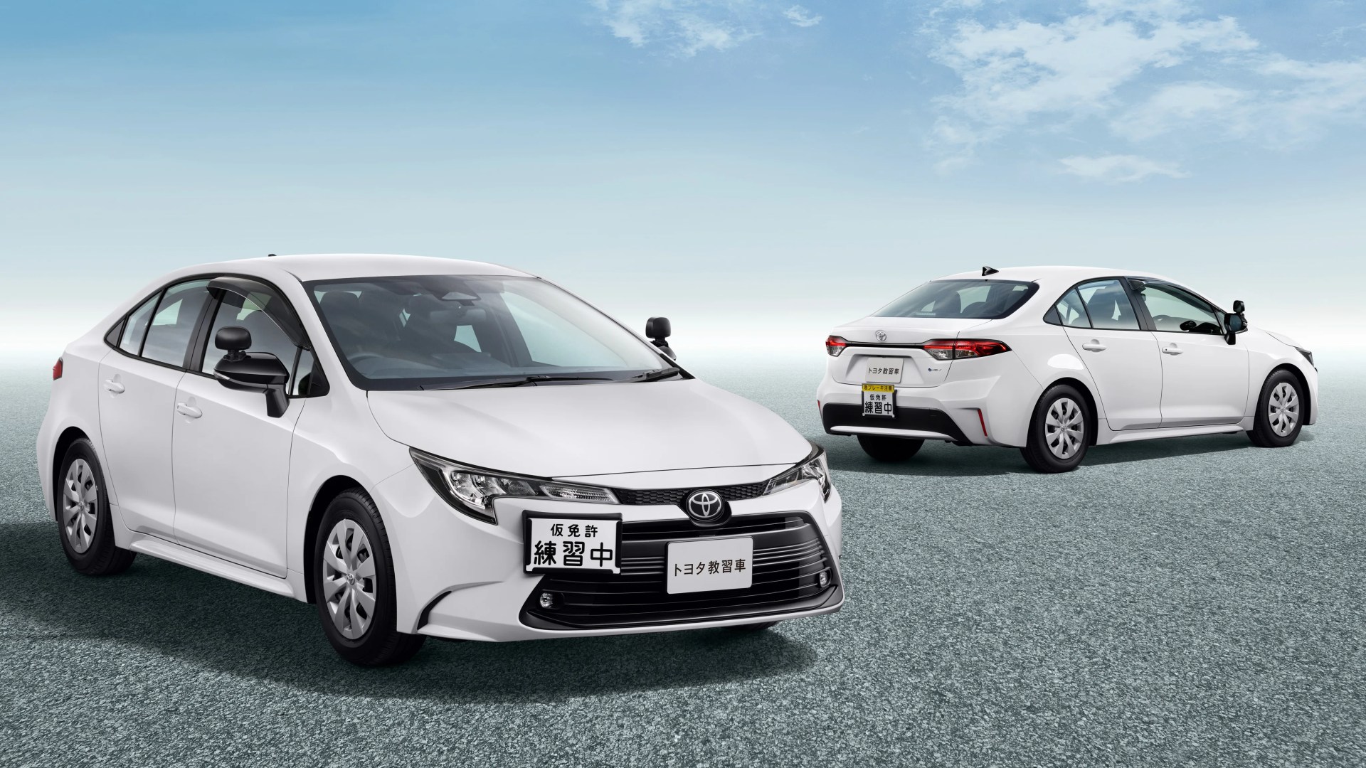 Two white Toyota sedans with Japanese learner driver signs on front and rear license plates, parked on a textured surface under a blue sky.