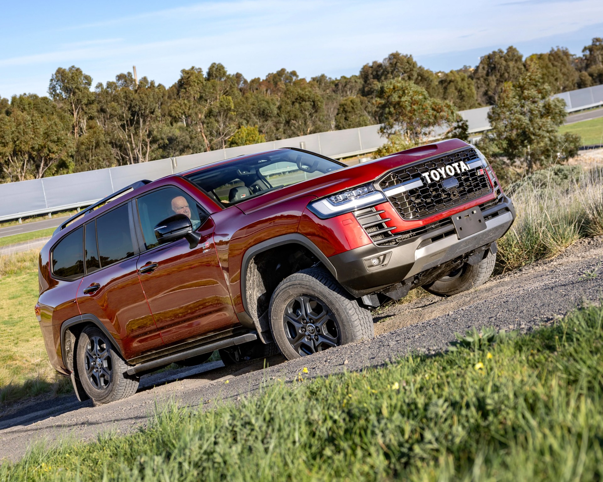 Red Toyota SUV with black wheels driving on a gravel road with trees in the background.