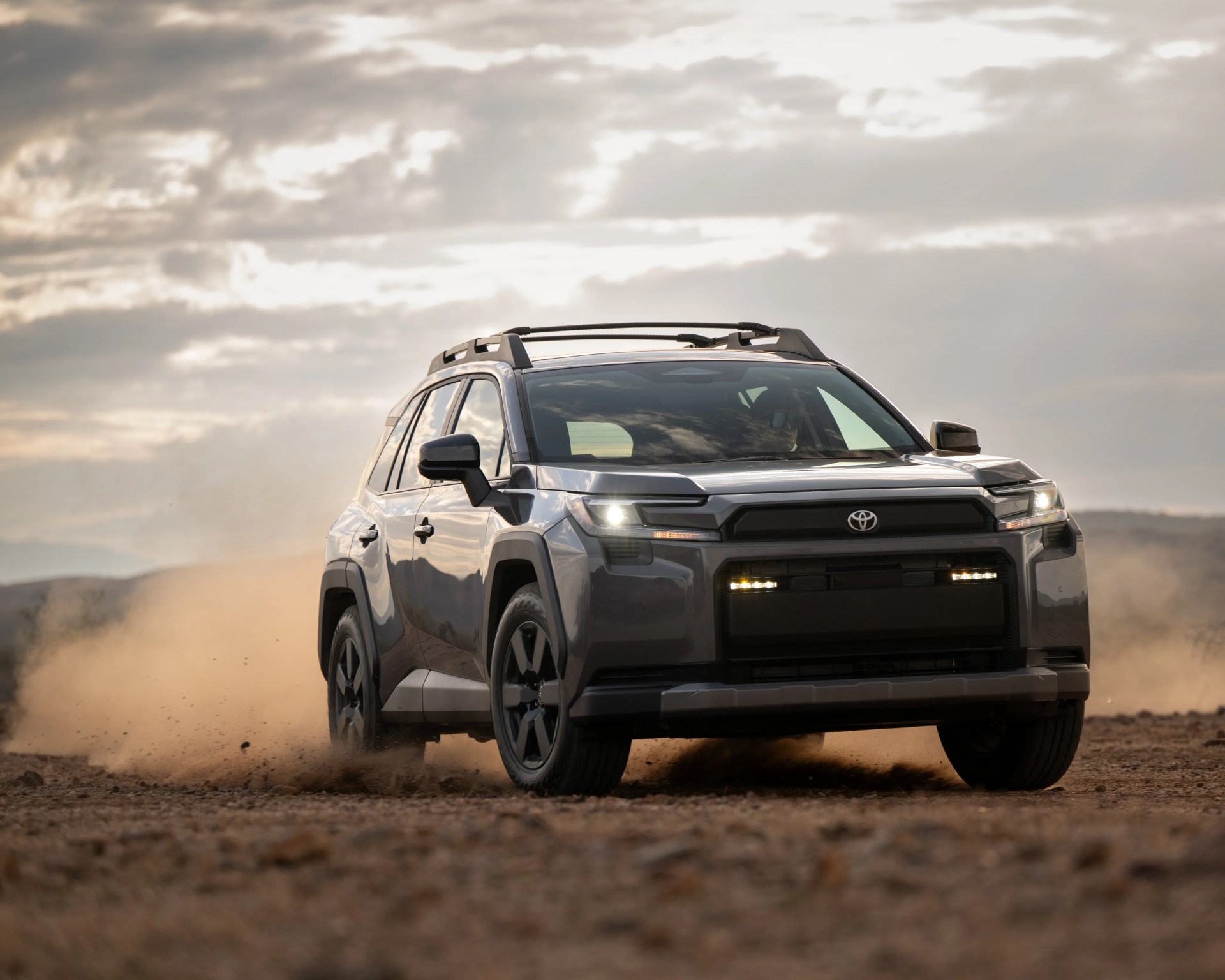 Gray Toyota SUV driving on a dusty dirt road with headlights on under a cloudy sky.
