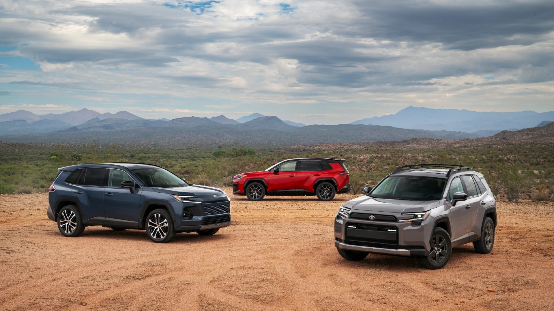Three Toyota RAV4 SUVs in gray, blue, and red parked on a dirt area with mountains and cloudy sky in the background.