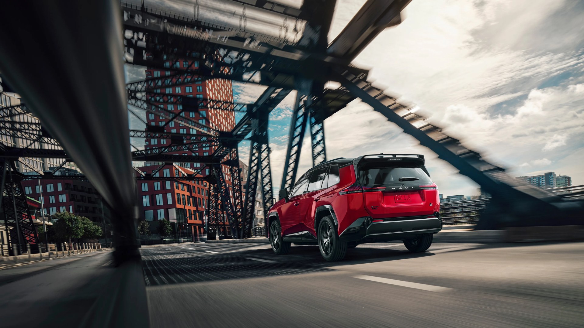 Red and black Toyota RAV4 SUV driving on a city bridge with steel beams and modern buildings in the background.