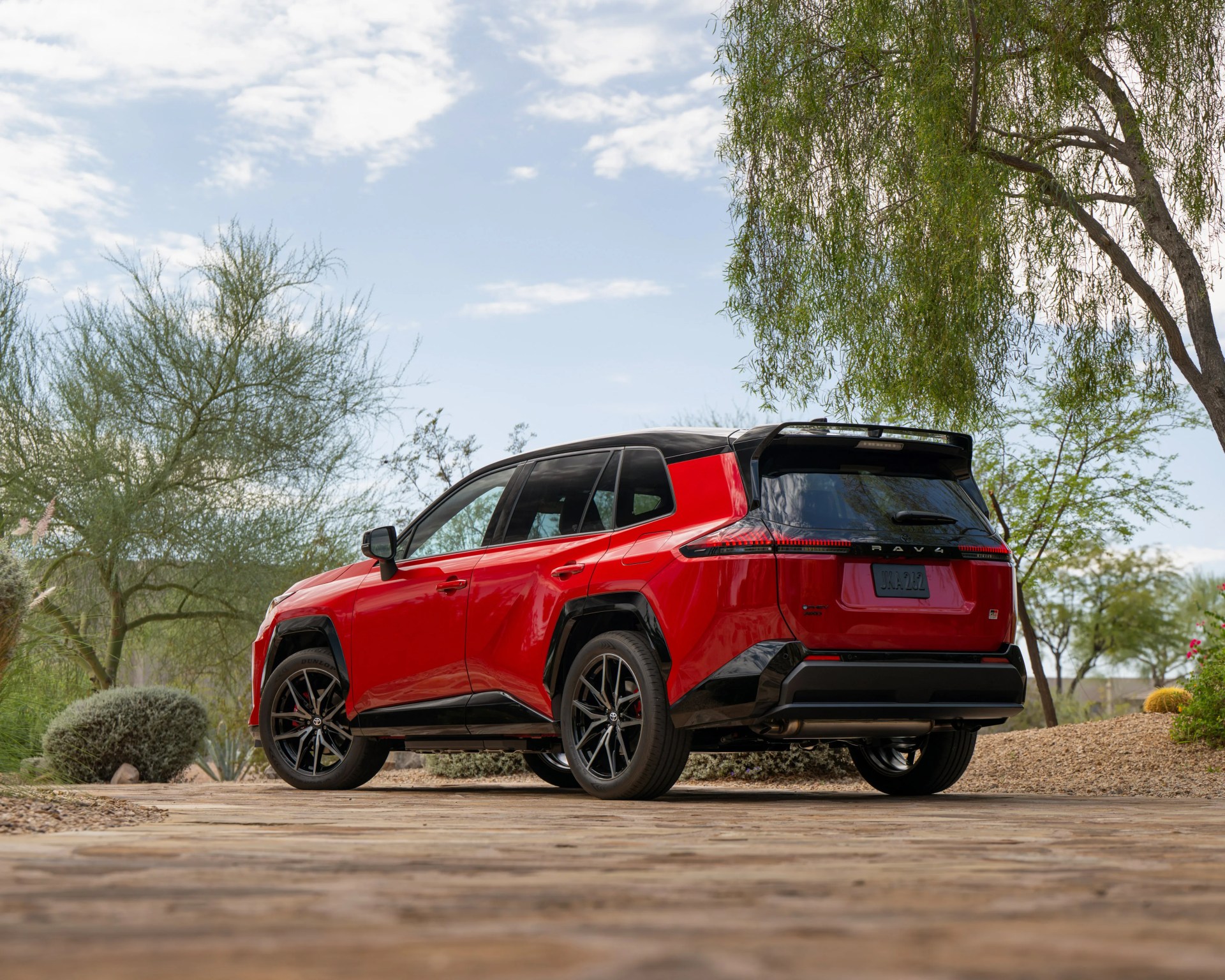 Red and black Toyota RAV4 parked on a stone driveway with desert plants and trees in the background.