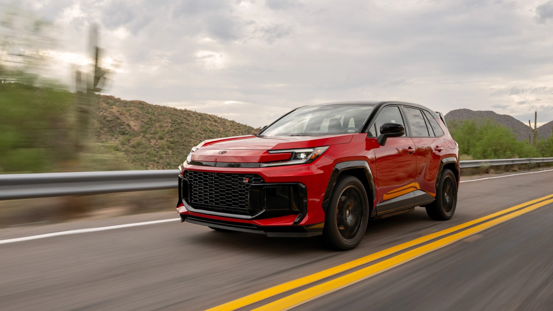 Red and black Toyota SUV driving on a desert highway with mountains and cloudy sky in the background.