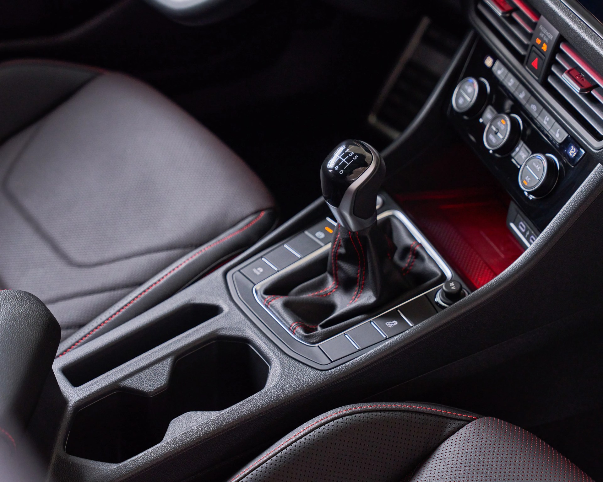 Black manual gear shift knob with six-speed pattern and black leather boot with red stitching in a car interior.