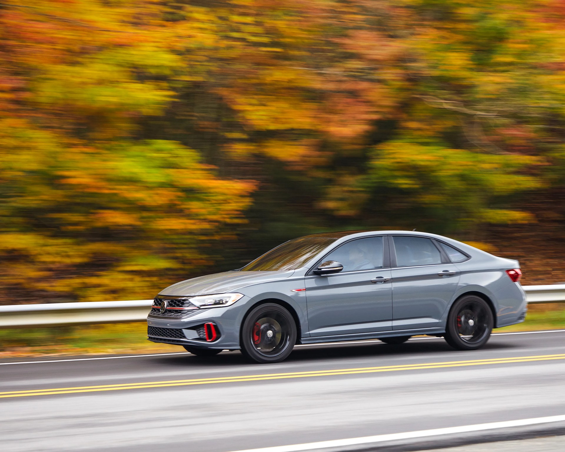 Gray Volkswagen sedan with black wheels driving on a road with autumn foliage in the background.
