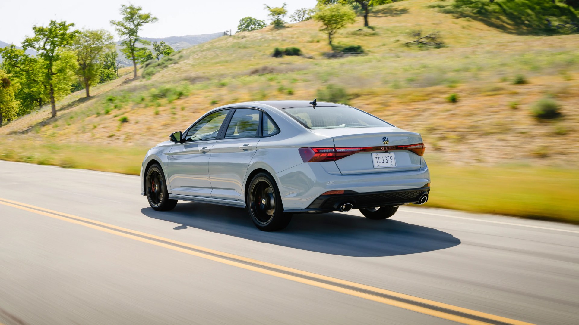 Silver Volkswagen GLI sedan with black wheels driving on a rural road with grassy hills in the background.
