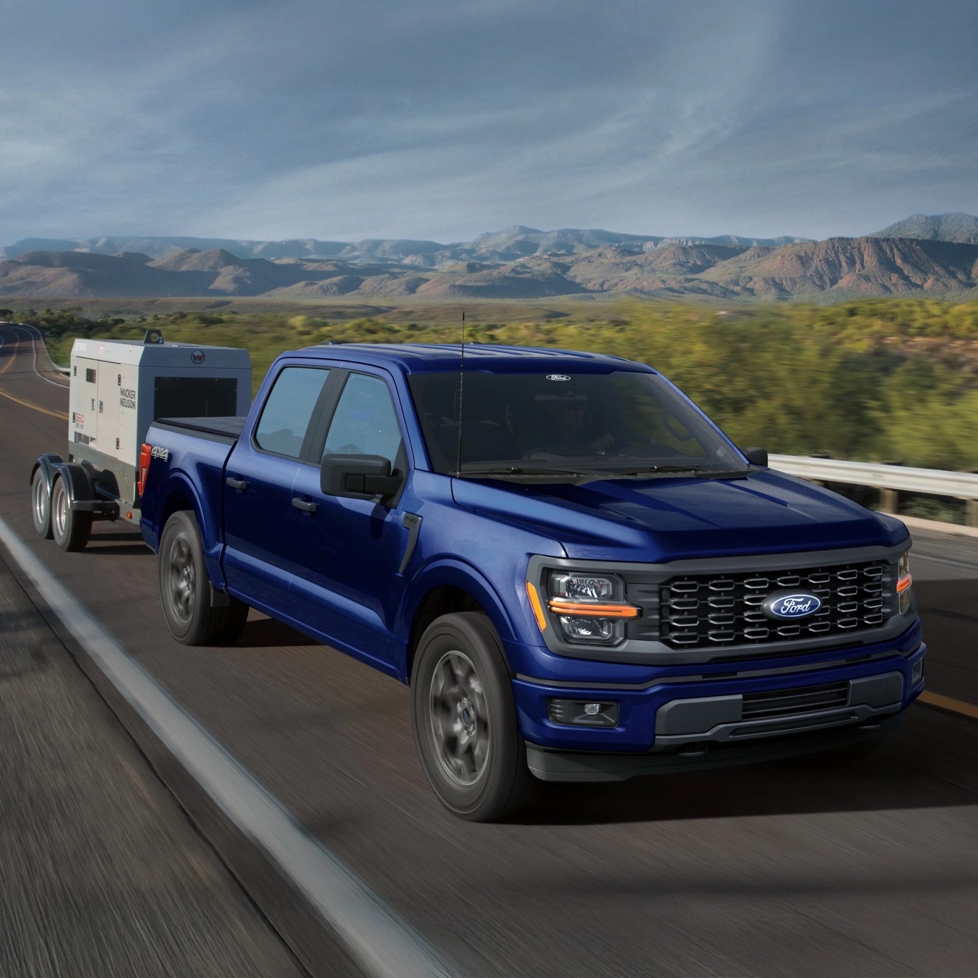 Blue Ford pickup truck towing a white Wacker Neuson generator on a highway with mountains in the background.
