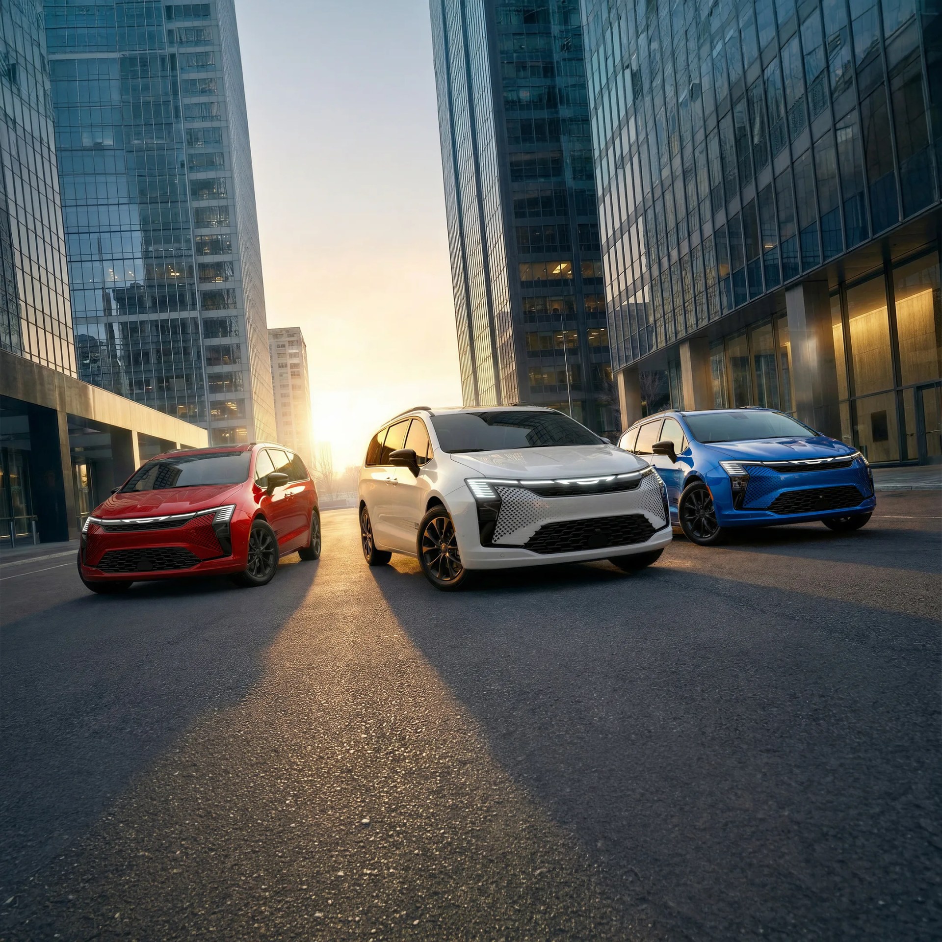 Three modern electric cars in red, white, and blue parked on a city street between tall glass buildings at sunset.
