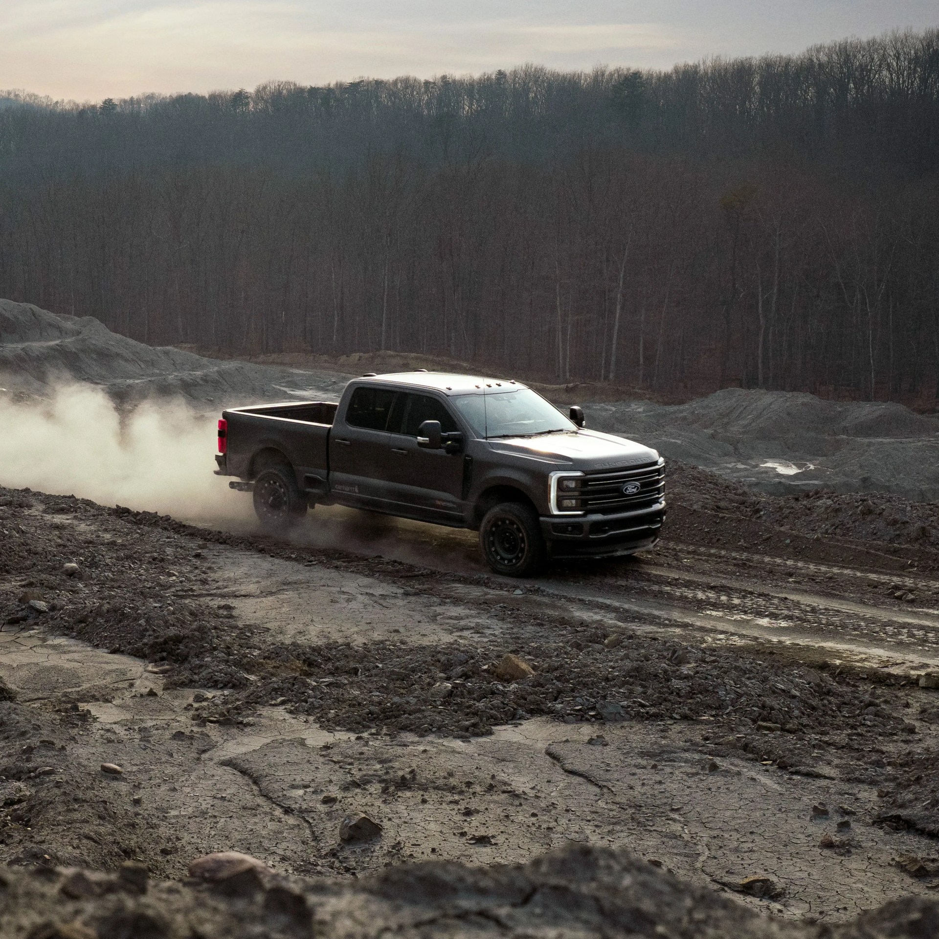 Black Ford Super Duty pickup truck driving on a dusty, rugged dirt road with bare trees in the background.