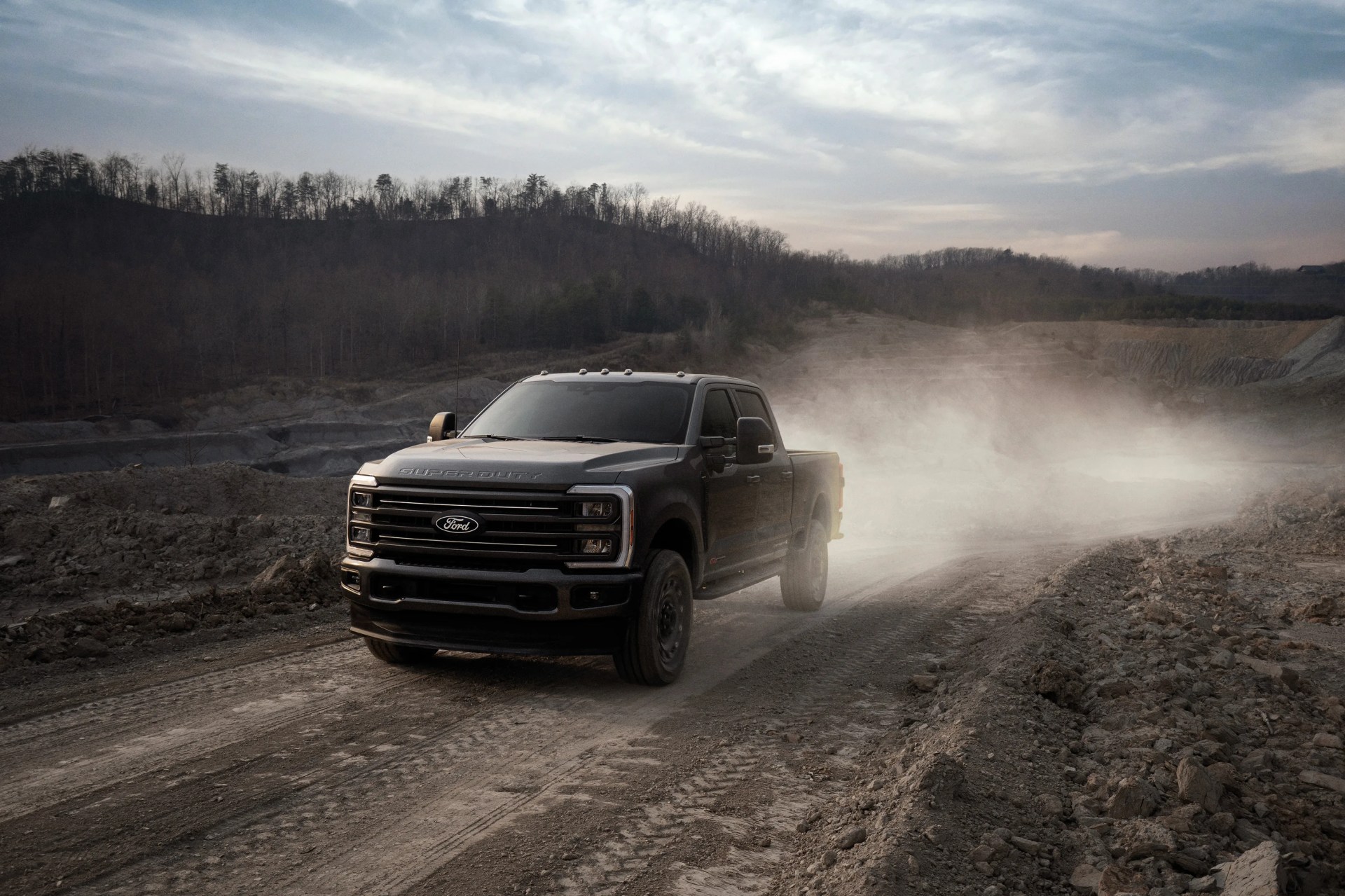 Black Ford Super Duty pickup truck driving on a dusty dirt road with hills and trees in the background.