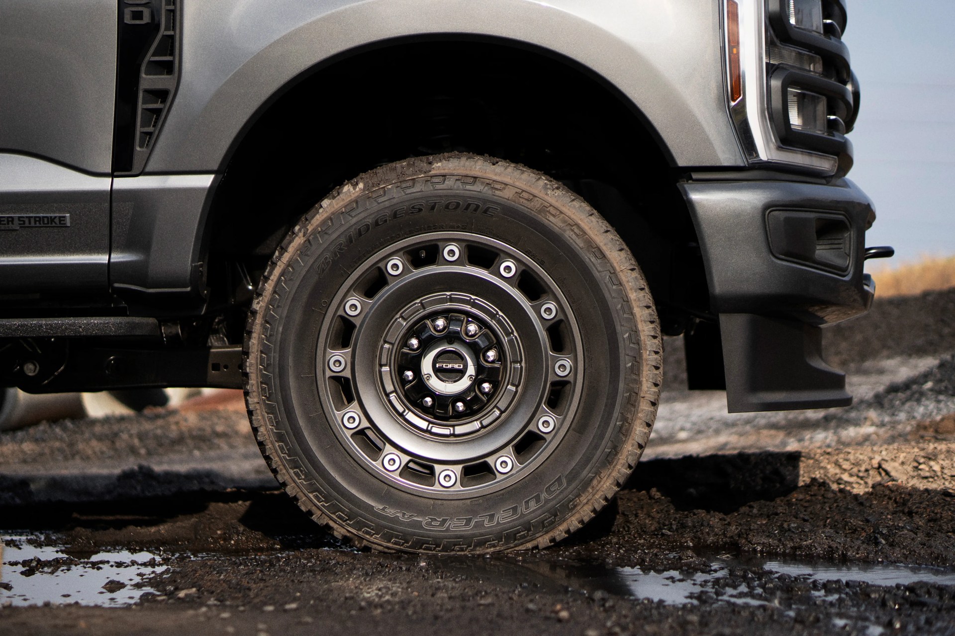 Close-up of a dirty Bridgestone Dueler A/T tire mounted on a black Ford truck wheel with a gray truck body above.