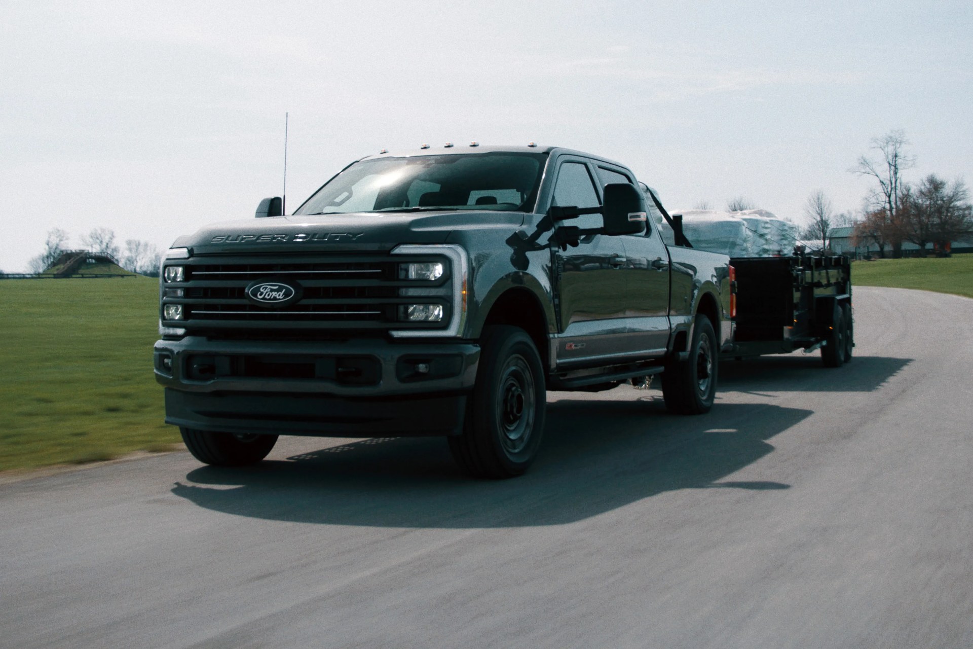 Dark gray Ford Super Duty pickup truck towing a trailer loaded with wrapped cargo on a paved road.