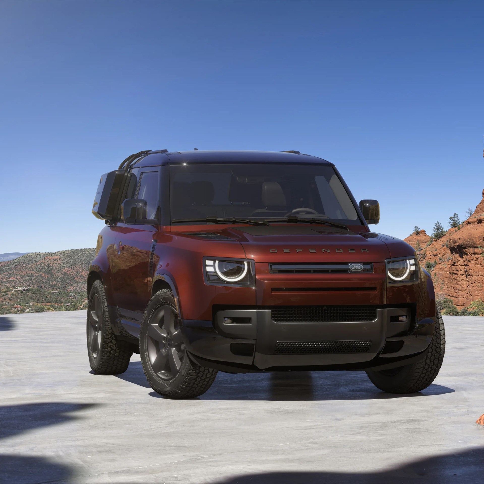 Red Land Rover Defender with black roof and rugged tires parked on concrete with rocky landscape in background.