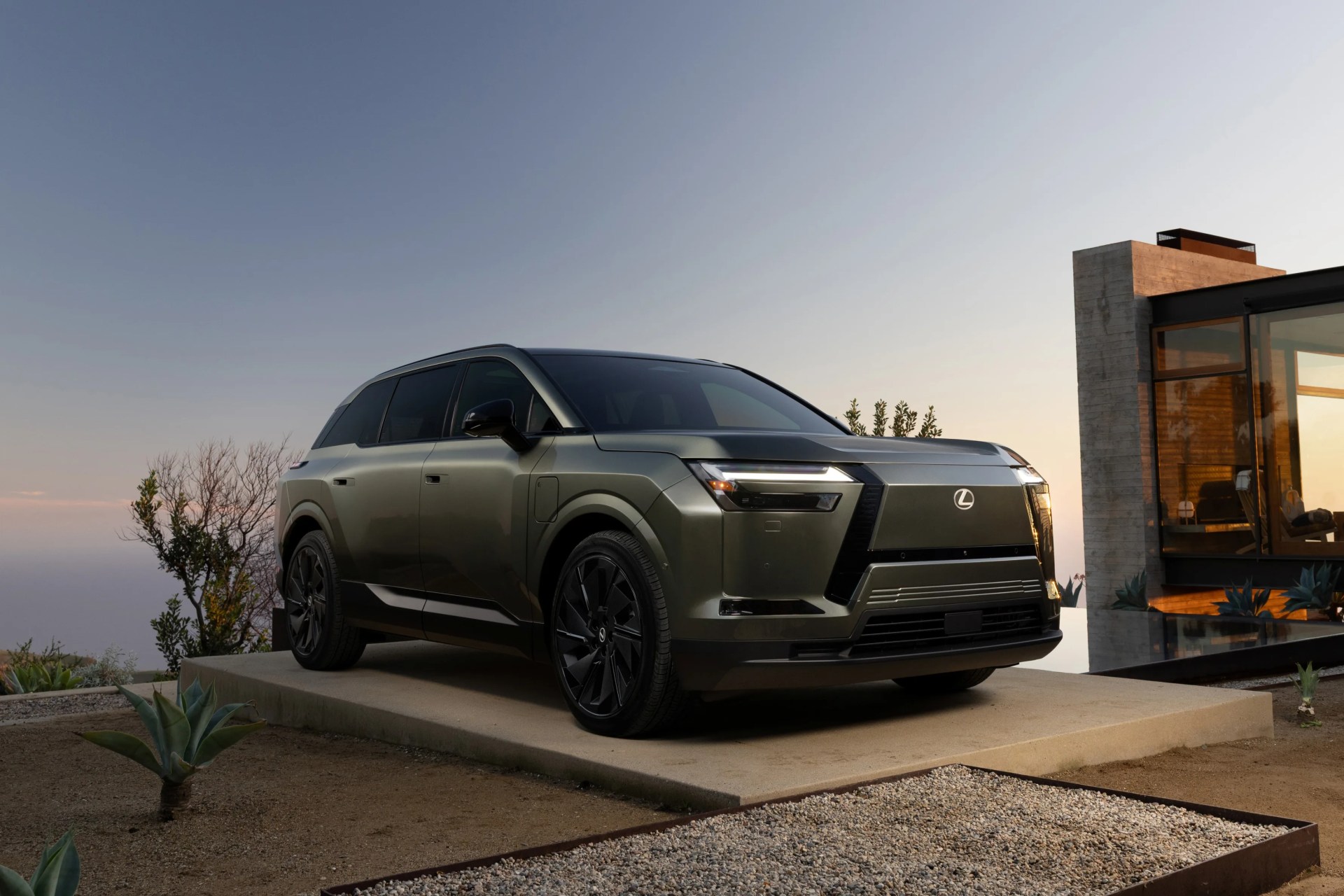 Matte green Lexus SUV with black wheels parked on a concrete platform near a modern glass and concrete building at sunset.