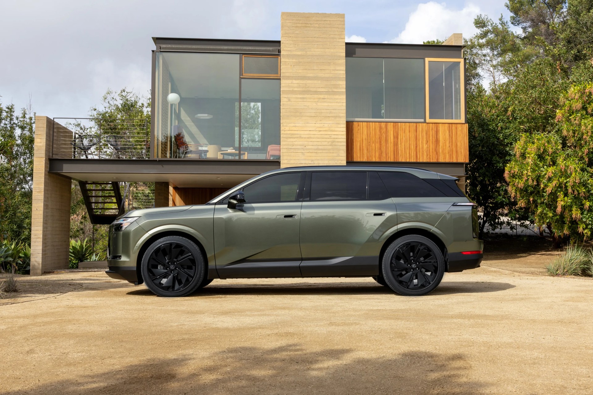 Green metallic SUV with black wheels parked in front of a modern house with large glass windows and wooden accents.