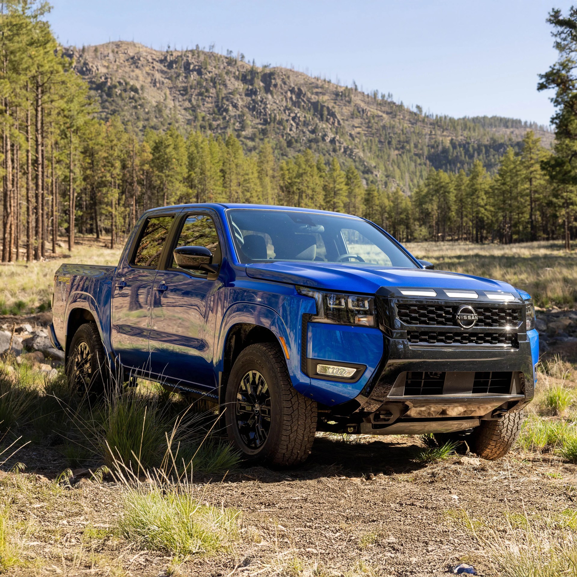 Blue Nissan pickup truck parked on a dirt path with forest and mountains in the background.