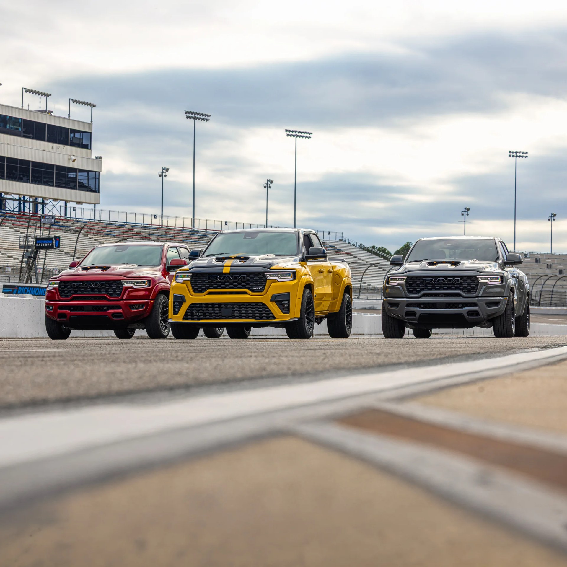 Three Ram pickup trucks in red, yellow with black stripes, and gray parked side by side on a racetrack.