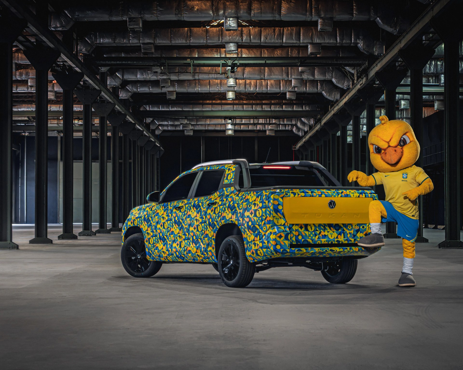 Blue and yellow patterned Volkswagen pickup truck with black wheels in a large industrial space next to a person in a yellow bird mascot costume wearing a Brazil soccer jersey.