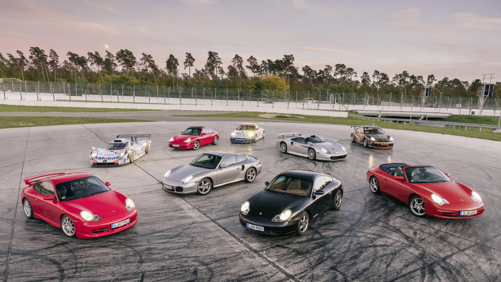Eight Porsche sports cars in various colors, including red, black, silver, and white, arranged on a racetrack circle with trees and fencing in the background.