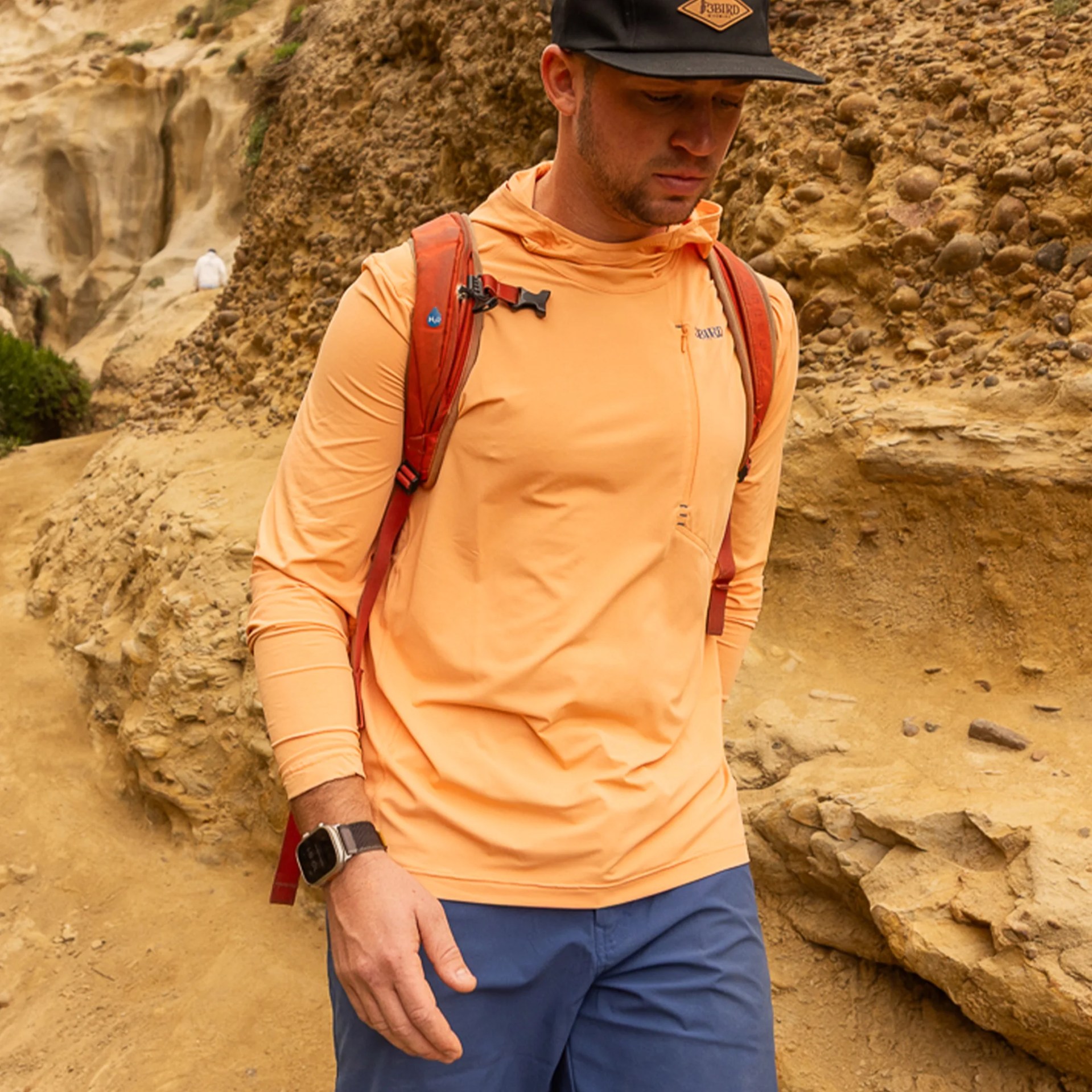 Man wearing a peach long-sleeve hoodie, red backpack, black cap, and blue pants standing on a rocky trail.