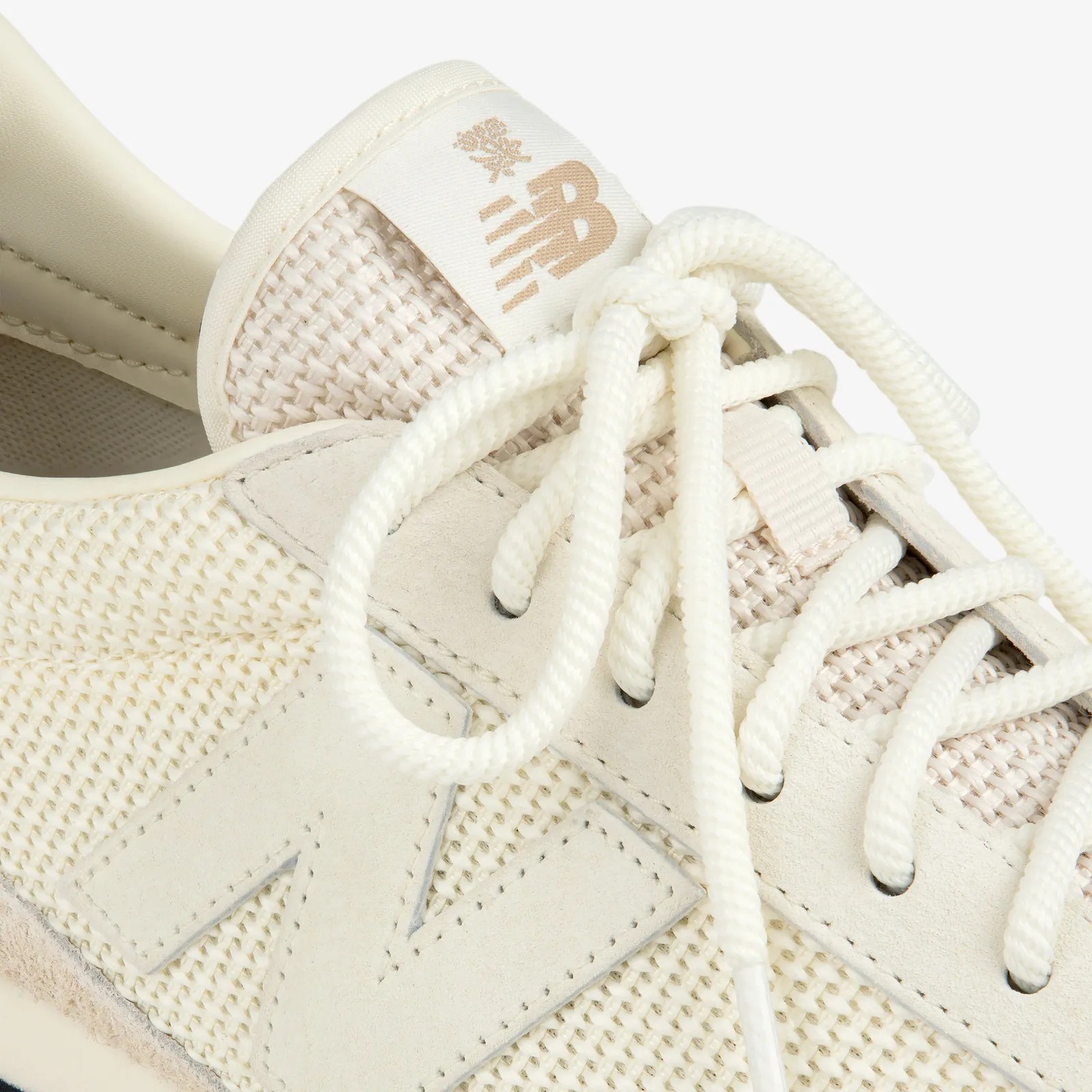 Close-up of beige New Balance sneaker with woven fabric, suede "N" logo, and thick white laces.