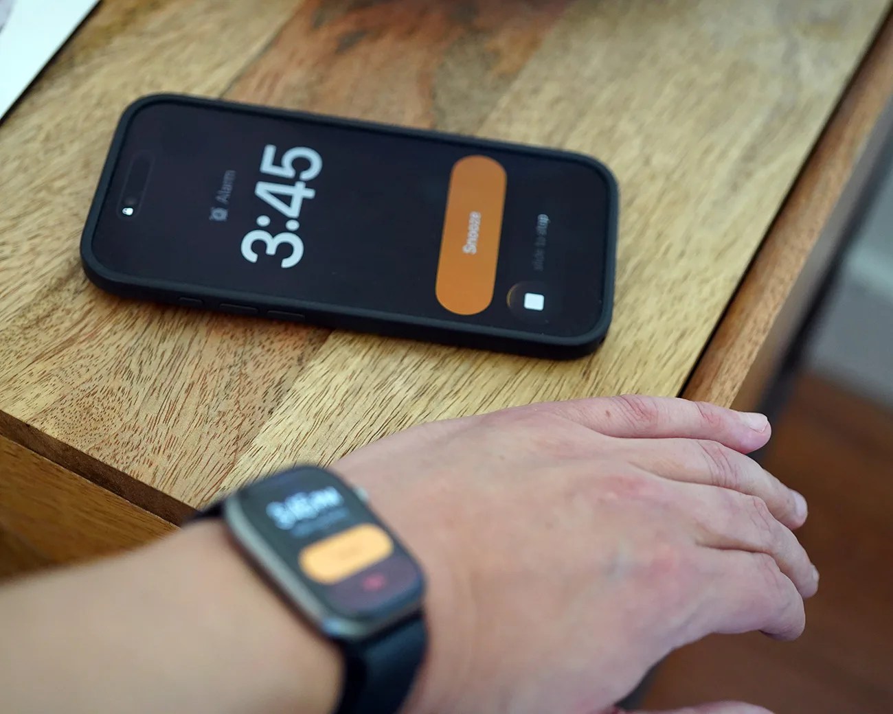 Black smartphone and smartwatch both displaying a 3:45 alarm snooze screen, with the phone on a wooden surface and the watch worn on a wrist.