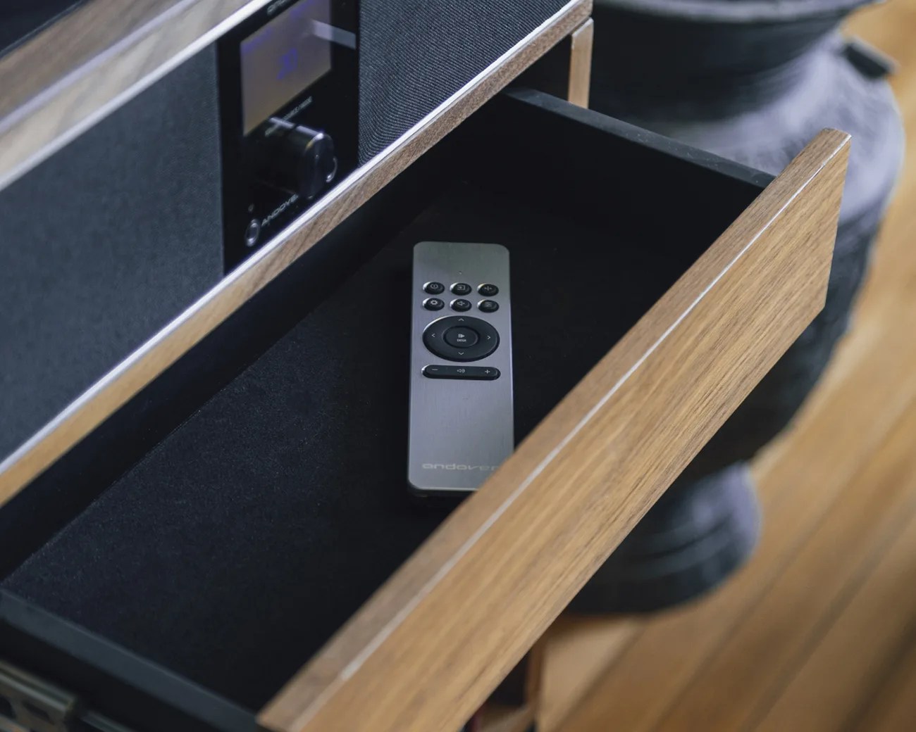Silver remote control with black buttons inside a wooden drawer lined with black fabric.