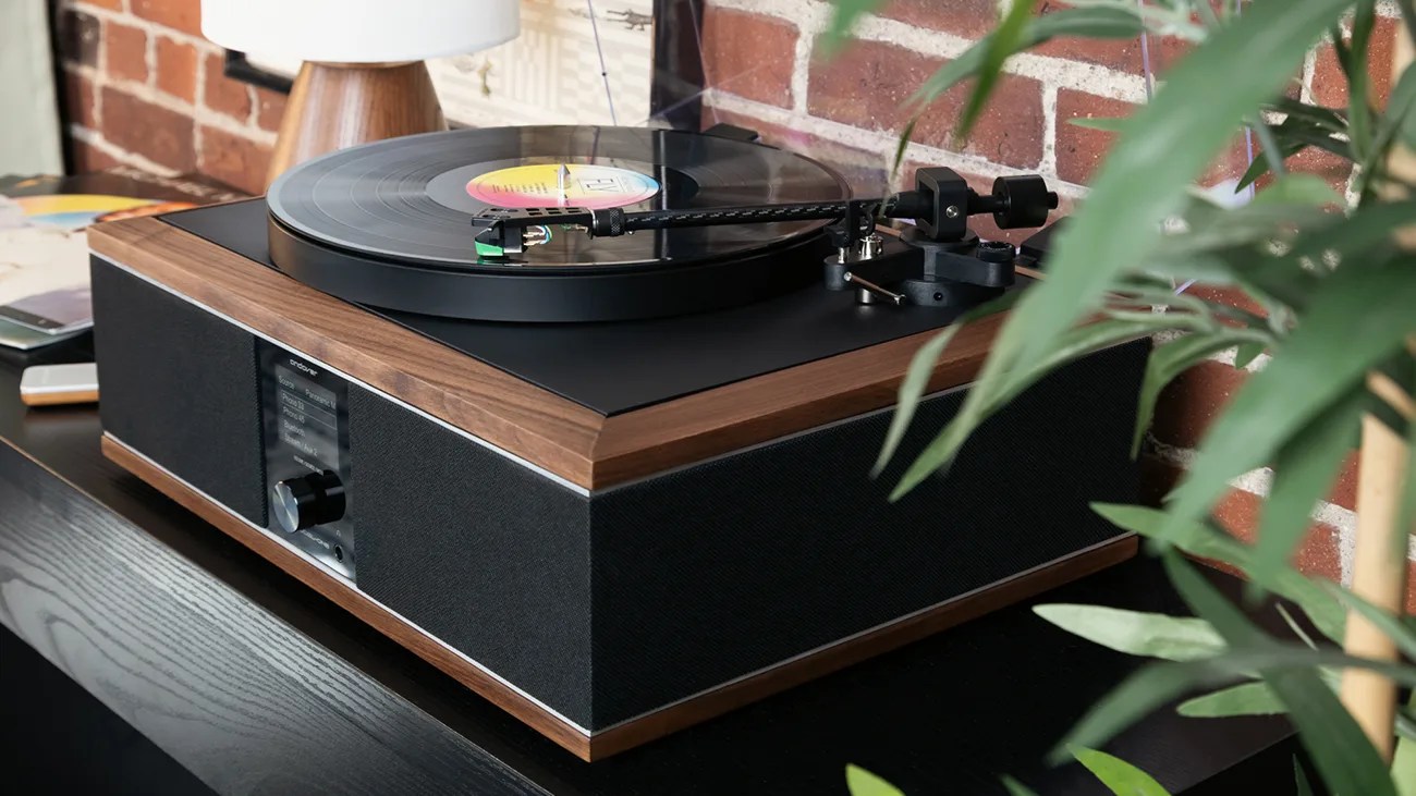 Wooden turntable with black speaker fabric and a vinyl record playing, placed on a black surface near a brick wall.