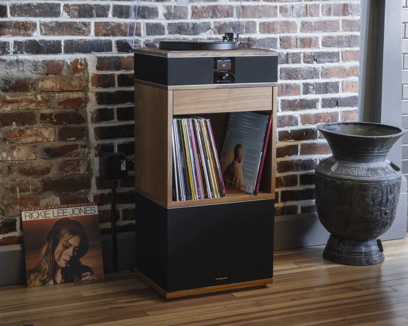 Wooden record player cabinet with black speaker panels, vinyl records inside, and a record leaning against a brick wall.