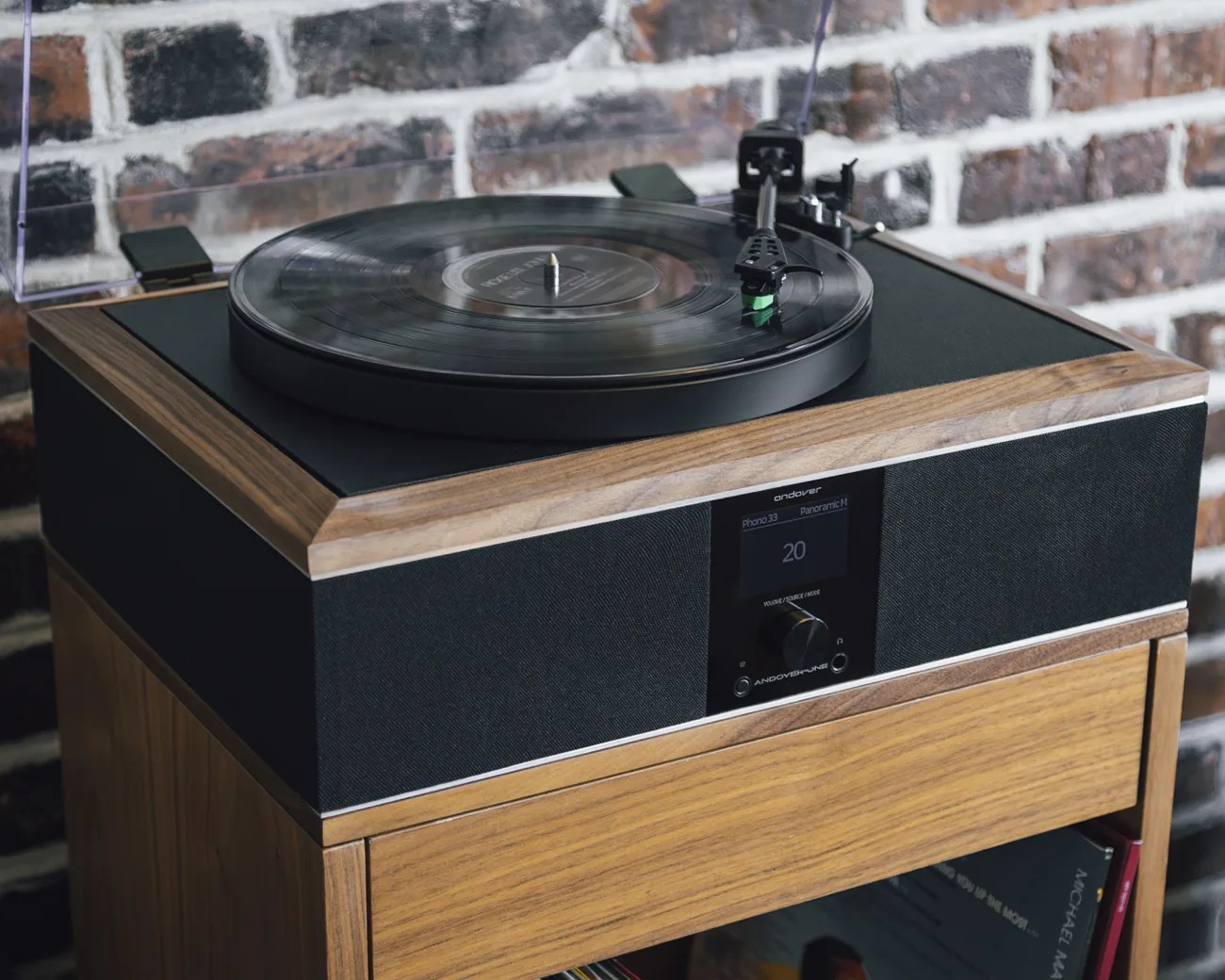 Wooden turntable with black speaker fabric and digital display on a wooden stand against a brick wall.