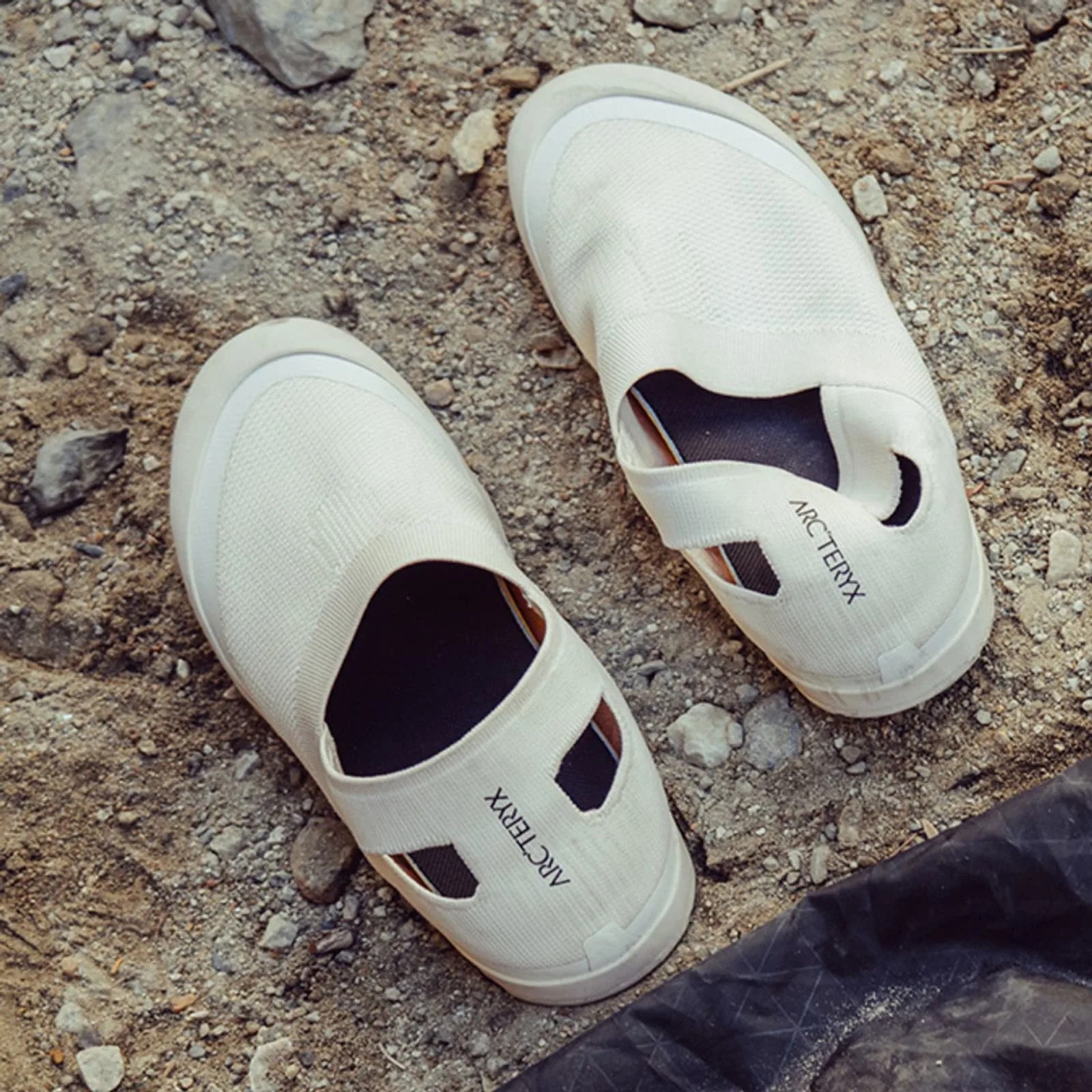 Pair of off-white slip-on shoes with black insoles and "ARC'TERYX" branding on a rocky ground.