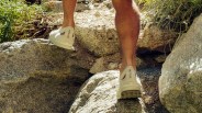 White Arc'teryx shoes worn by a person walking on rocky terrain with green plants nearby.