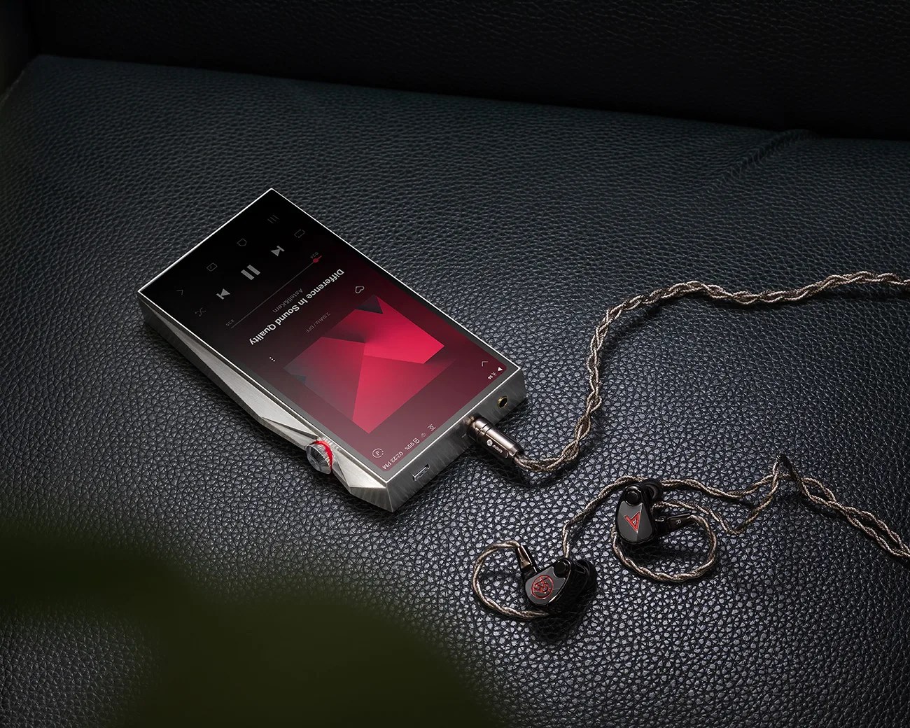 Silver digital music player with a red volume knob connected to black in-ear monitors with braided cables on a black leather surface.