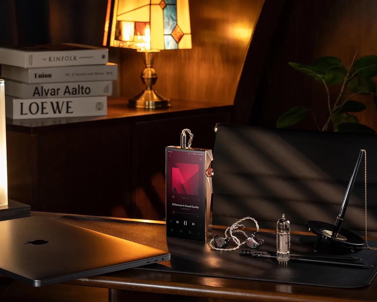 Metallic digital audio player with earphones and vacuum tube on a leather desk mat next to a closed laptop and pen stand.